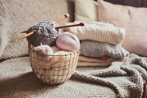 Ball of yarn with knitting needles sits in a basket on top of a handmade blanket.